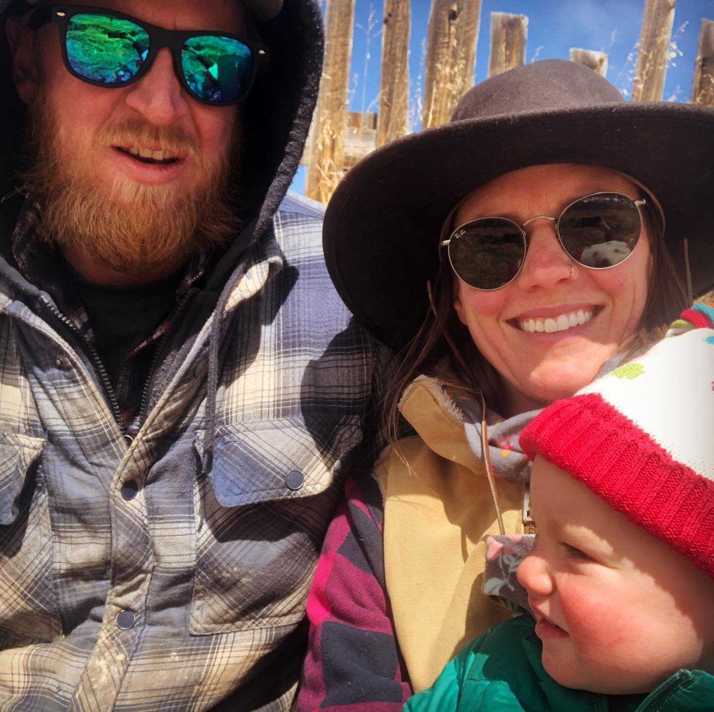 Family portrait featuring a man with a beard and sunglasses, a woman in a wide-brimmed hat and sunglasses, and a young child wearing a colorful knit hat. The background shows a wooden fence and blue sky, capturing a joyful outdoor moment.