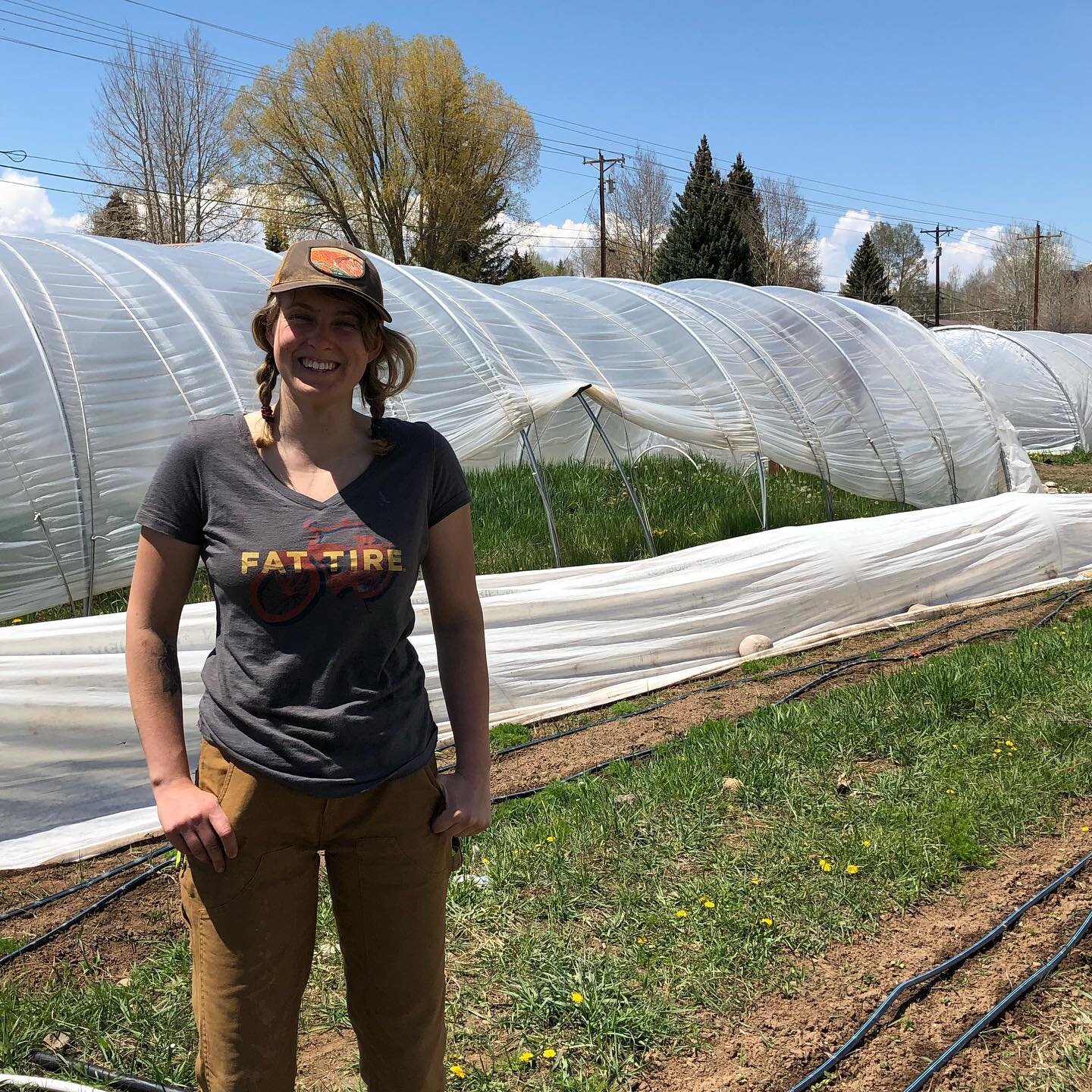 Young woman standing in a sunny farm setting, wearing a "Fat Tire" t-shirt and brown pants, with green grass and plastic greenhouses in the background. Ideal for content related to sustainable farming, agriculture, or outdoor lifestyle.