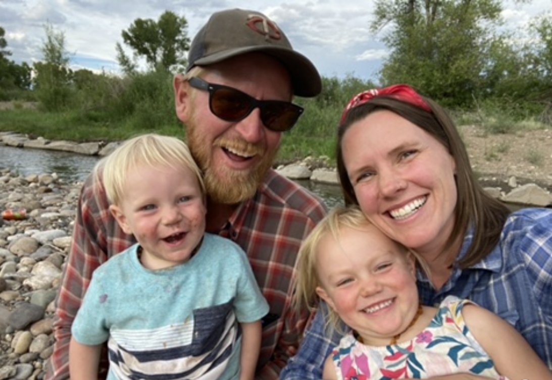 IMG-1391 Family portrait by a river featuring two smiling children and their parents, enjoying a sunny day outdoors. The scene captures a joyful moment with natural scenery in the background.